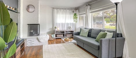 A living room with a grey sofa, a white rug, a coffee table, a fireplace, and a large window with curtains.