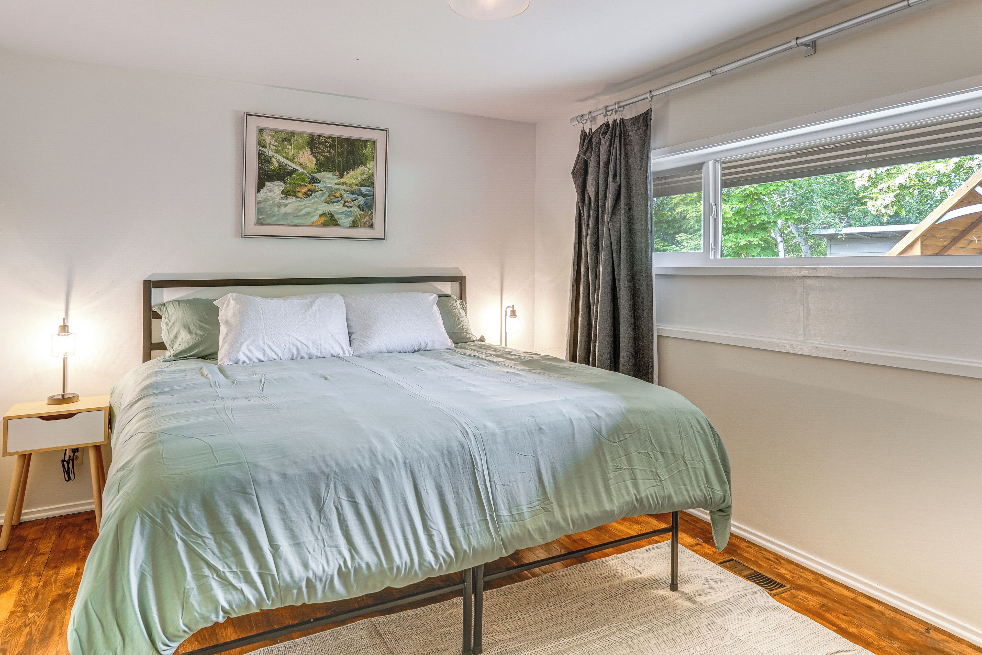 Une chambre à coucher avec un lit, une table de chevet, une lampe, une fenêtre avec des rideaux et un tableau au mur.