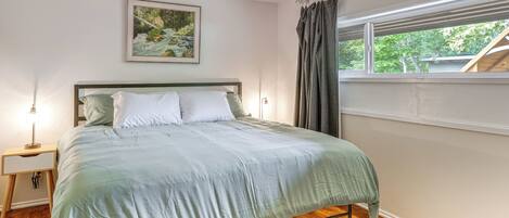 Une chambre à coucher avec un lit, une table de chevet, une lampe, une fenêtre avec des rideaux et un tableau au mur.