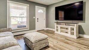A living room with a large flat-screen TV mounted on the wall, a white entertainment center, a grey sofa, and a window with blinds. - Explore Downtown & Mass Ave! 3BR Indy Townhome (Indianapolis)