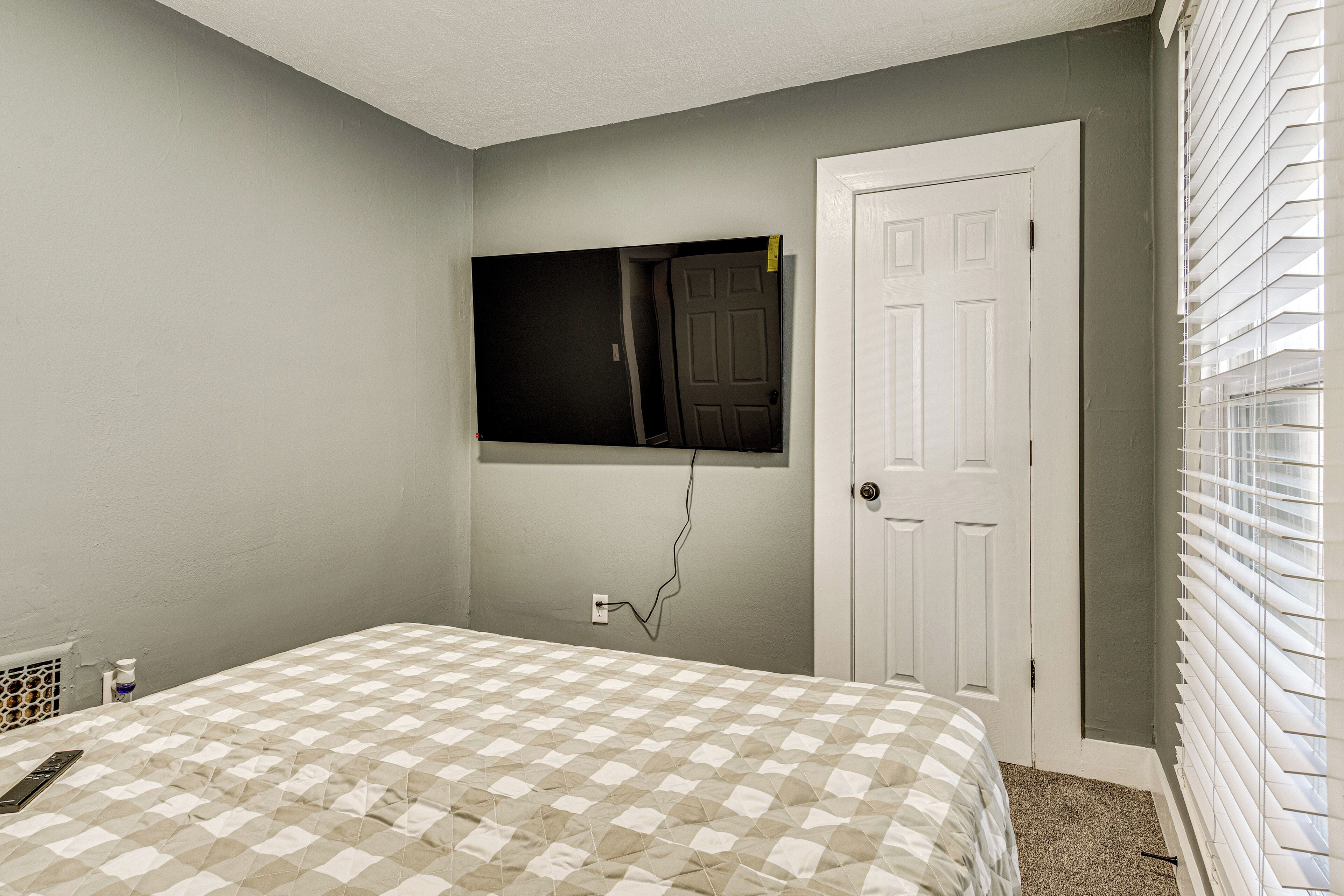 Une chambre à coucher avec une couvre-lit à carreaux, un téléviseur fixé au mur, une porte blanche et une fenêtre avec des stores.