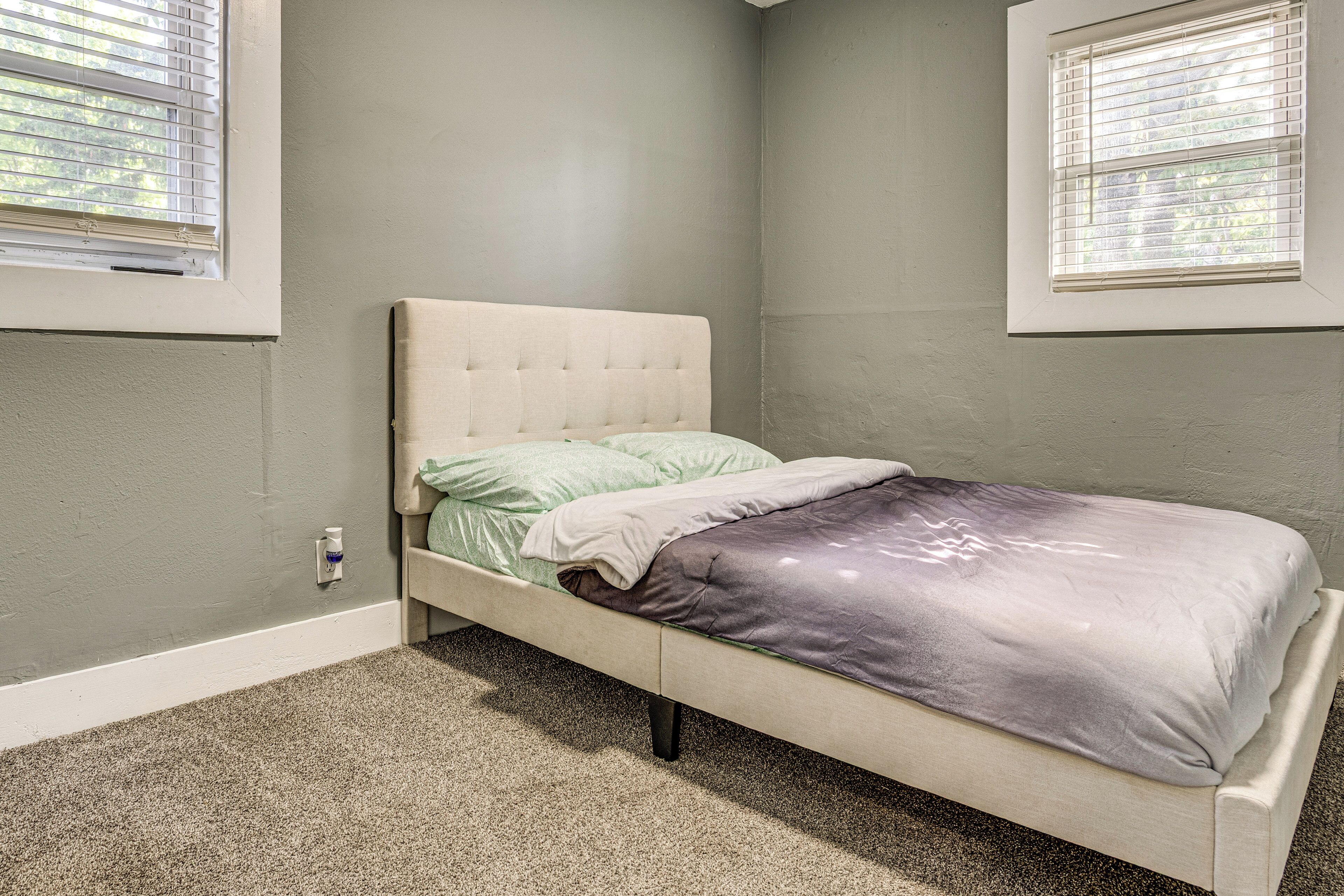 A bedroom with a single bed, a carpeted floor, and two windows with blinds.