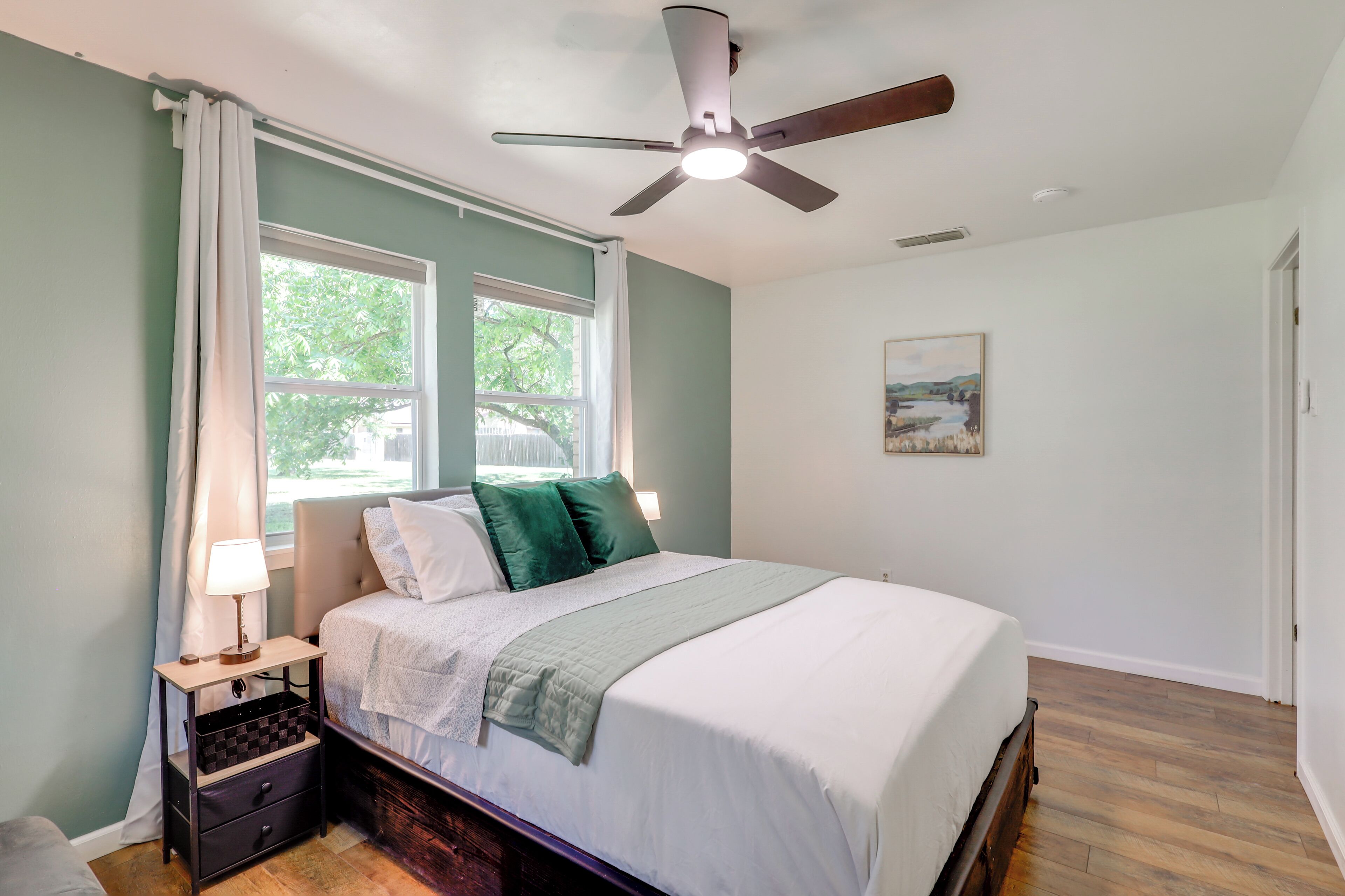 A bedroom with a bed, a nightstand, a ceiling fan, a window with curtains, and a painting on the wall.