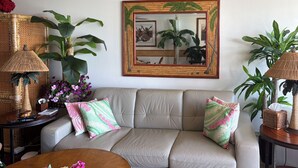 A living room with a beige sofa, two patterned pillows, a wooden coffee table with a floral arrangement, and large green plants. - Resort Condo on Oahu: Ocean & Pool Views! (Hau`ula)