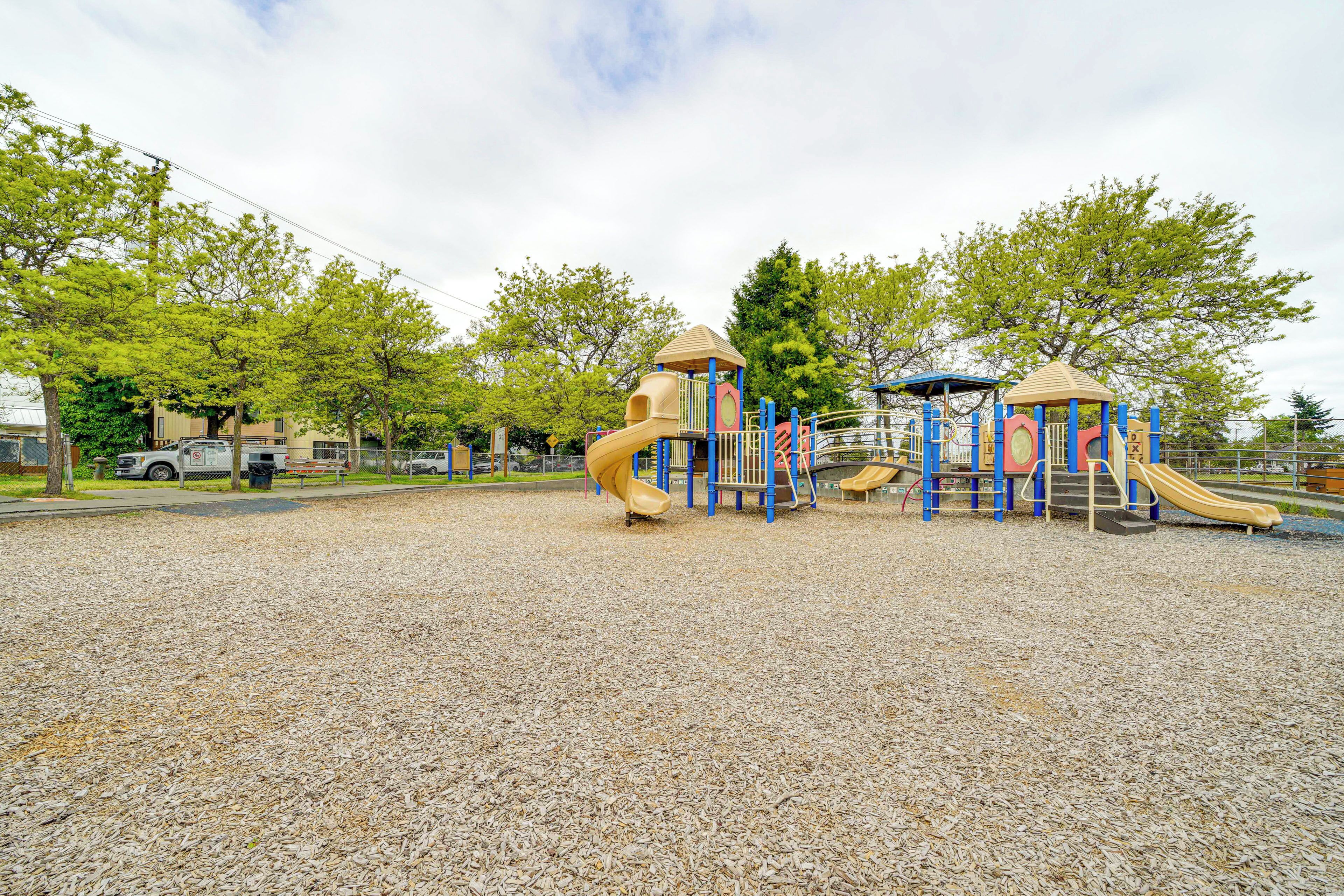 A playground with multiple slides and climbing structures, surrounded by trees and a gravel surface.