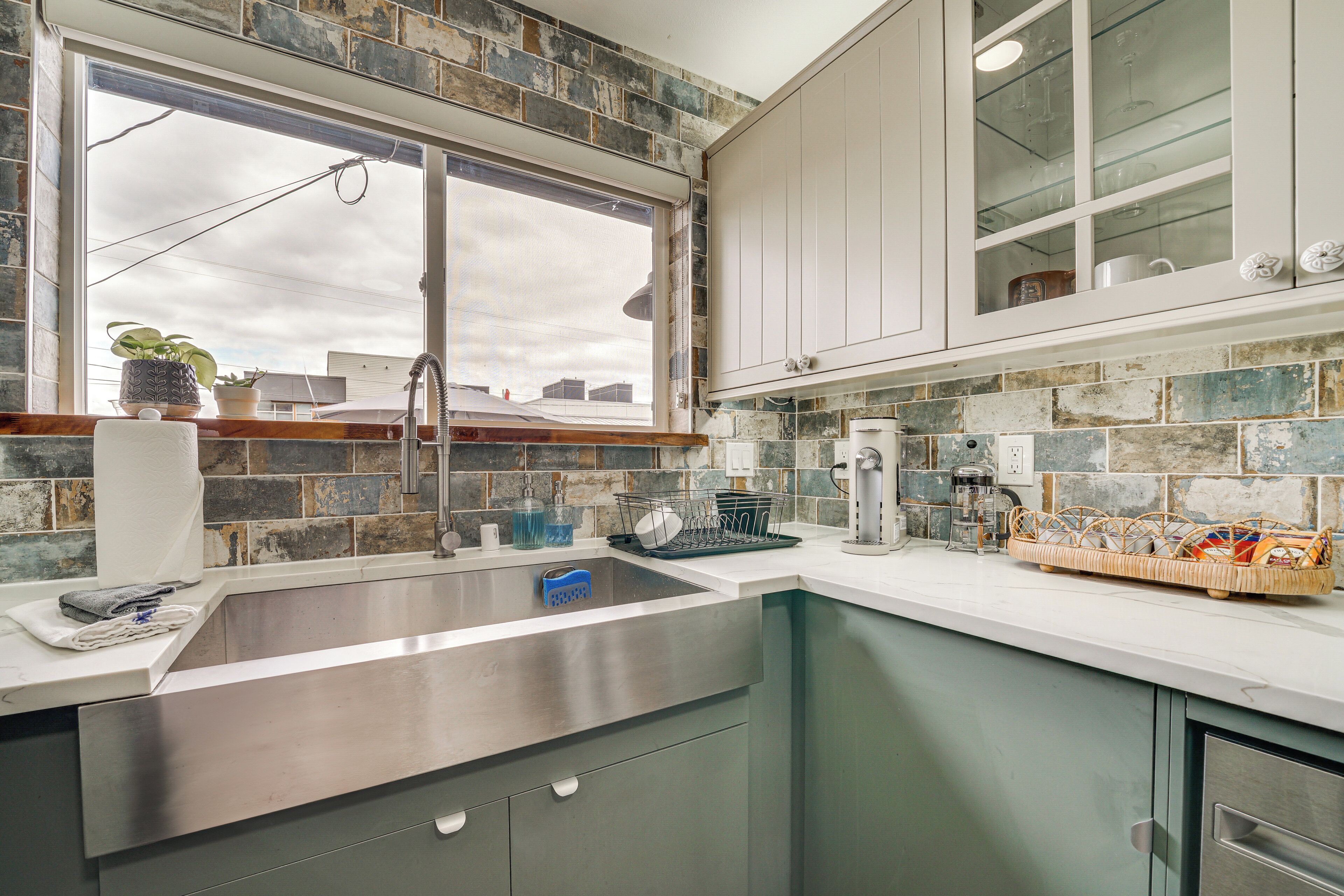 Una cocina con un lavamanos de acero inoxidable, armarios blancos y una ventana con vistas a edificios.
