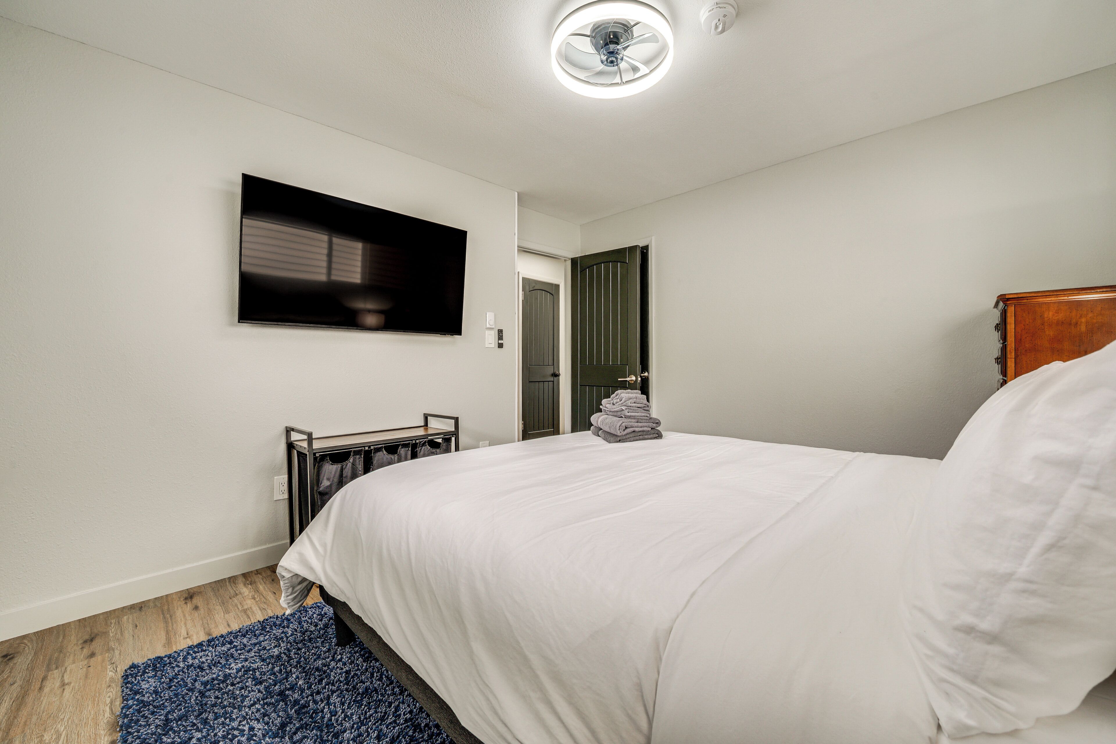 A bedroom with a bed, a TV mounted on the wall, a wooden floor, and a door leading to another room.