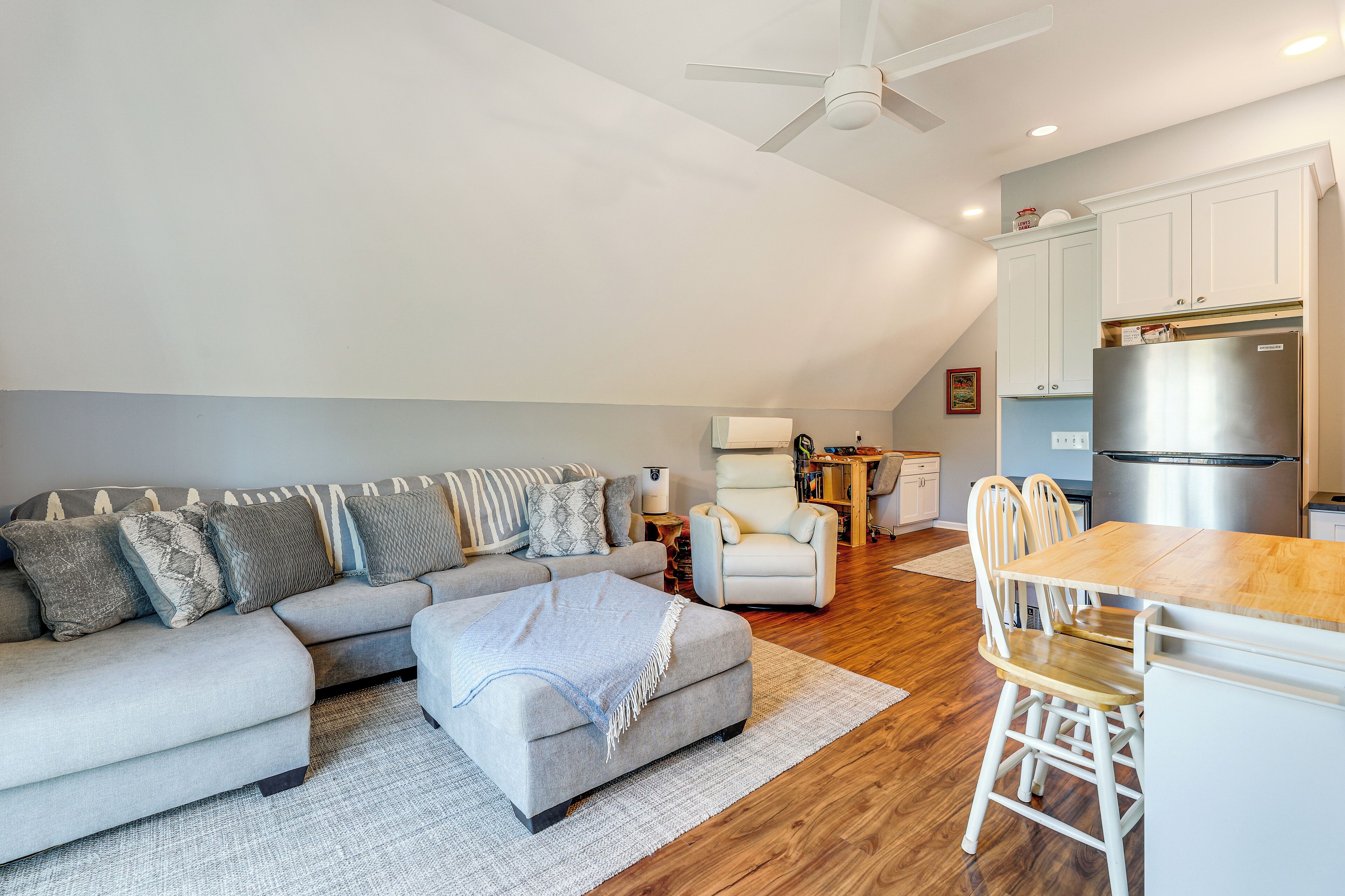 A modern living room with a sectional sofa, a wooden dining table, and a kitchen area with white cabinets and a stainless steel refrigerator.