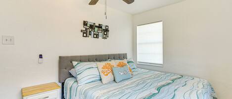Un dormitorio con cama, ventilador de techo, una obra de arte en la pared, mesita de noche y ventana.