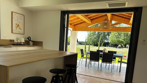 Une cuisine moderne avec un plan de travail en bois, des tabourets de bar et une vue sur une terrasse extérieure équipée d’une table et de chaises.