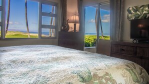 Una camera da letto con un letto grande, un comodino con una lampada, una TV su una cassettiera e vista sulla spiaggia dalle finestre.