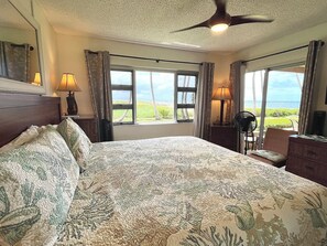 Une chambre d’hôtel avec un grand lit, deux fenêtres avec des rideaux, un ventilateur de plafond et une vue sur la mer.