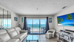 A modern living room with a sofa, coffee table, and TV, overlooking a balcony with a view of the beach.