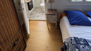 Un dormitorio con cama, mesita de noche, lámpara, ventana con vistas y puerta que da a otra habitación.