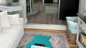 Una sala de estar con un sofá, una mesa de centro y un mueble para televisión. Se ve una cocina a través de una puerta abierta.
