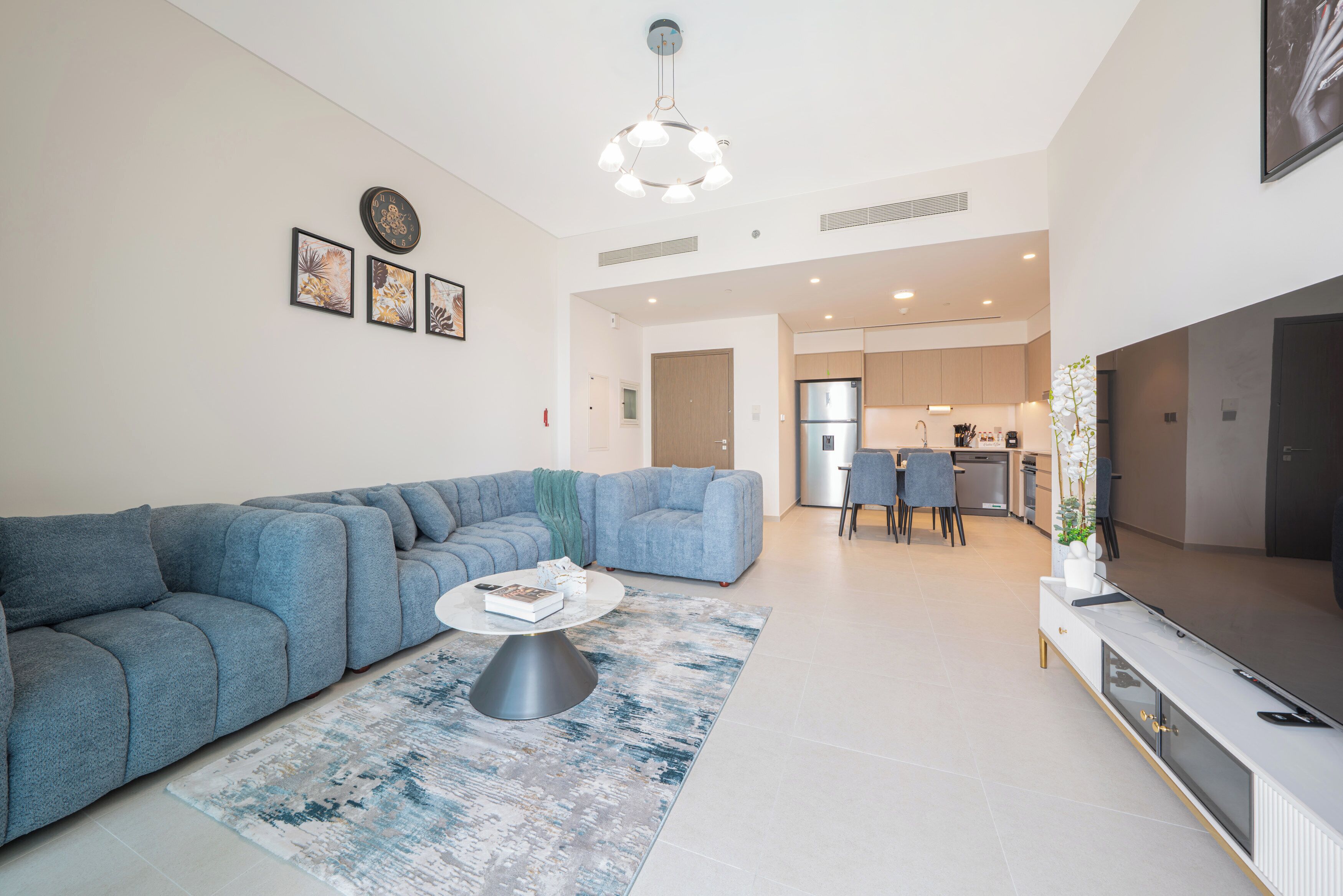 A modern living room with a blue sofa, a coffee table, and a TV. There is a kitchen in the background with a dining area.