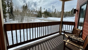 A wooden deck with a railing, a bench, and a view of a snowy landscape with trees and a road. - Unit 223 - Updated Condo (Winter Park)