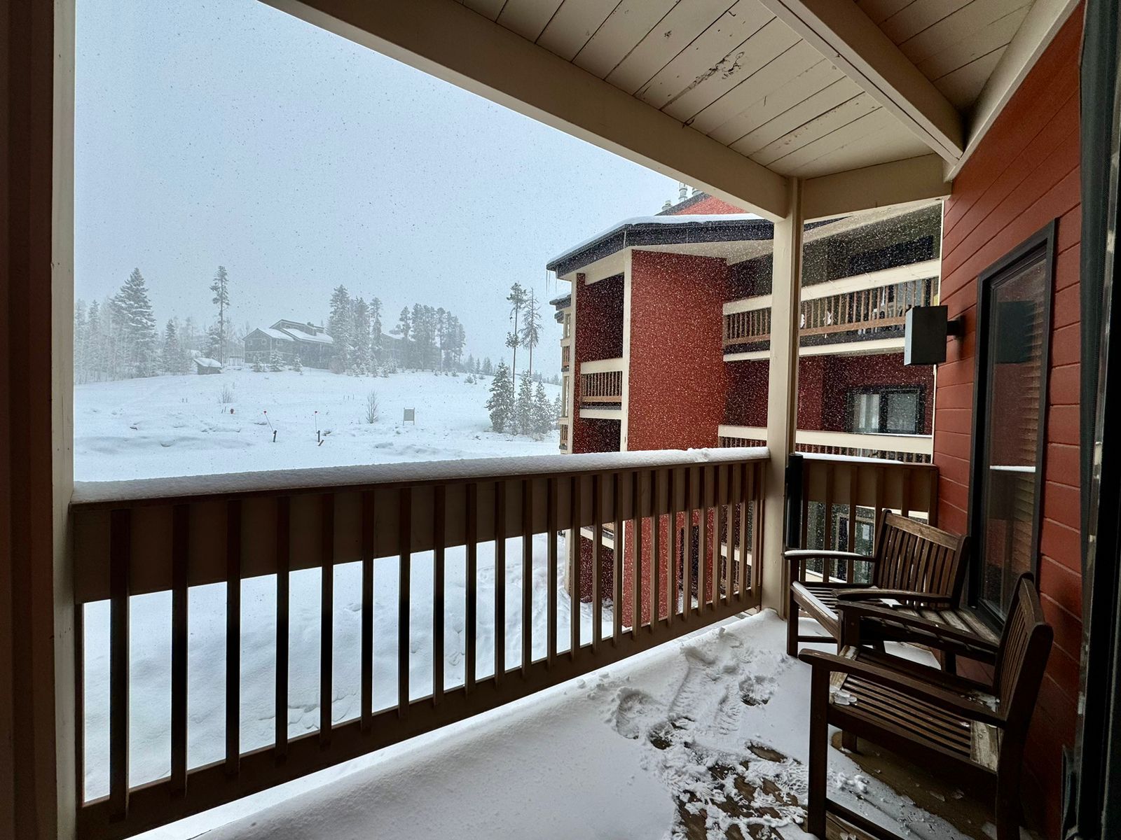 A covered balcony with a wooden railing, a bench, and a view of a snow-covered landscape with buildings in the background.