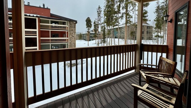 Une terrasse en bois avec une rambarde, deux chaises en bois et une vue sur un paysage enneigé, avec des bâtiments et des arbres.