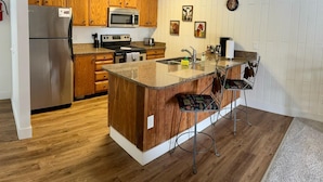 Une cuisine avec des meubles de rangement en bois, un réfrigérateur en acier inoxydable, un four à micro-ondes, une cuisinière et un évier. On y trouve deux tabourets de bar et une horloge au mur.