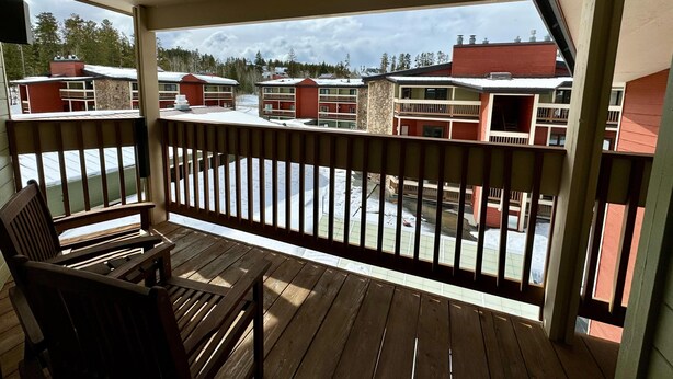 Een houten balkon met uitzicht op een rij rode en beige gebouwen, waarvan sommige ook balkons hebben, onder een heldere lucht.