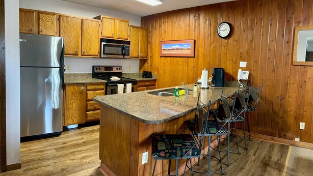 A kitchen with wooden cabinets, a stainless steel refrigerator, a microwave, an oven, and a sink. There are bar stools and a clock on the wall. - Unit 513 - Standard Condo (Winter Park)