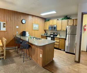 A kitchen with wooden cabinets, a stainless steel refrigerator, a microwave, a sink, and a bar stool.