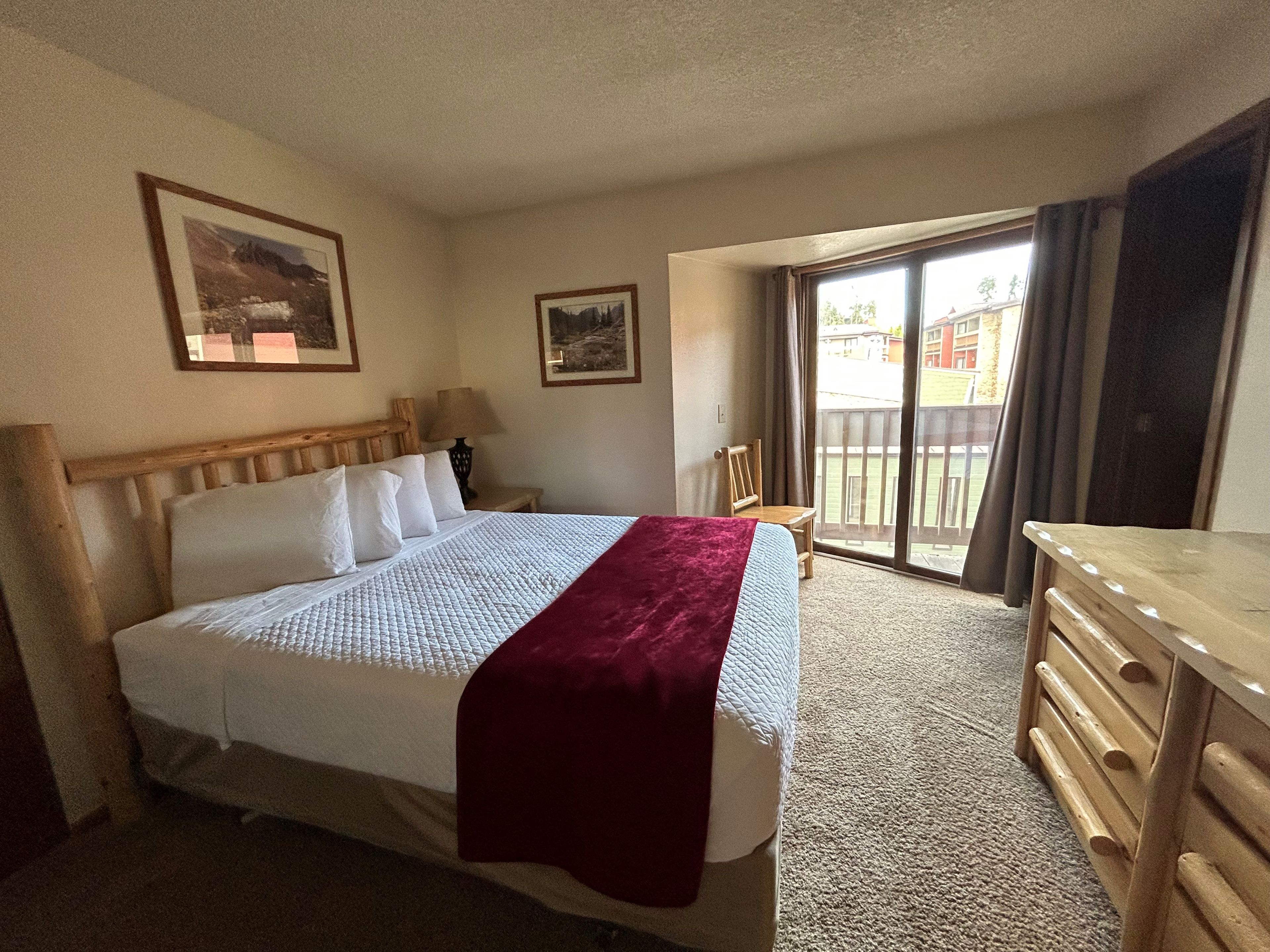 A hotel room with a bed, a wooden headboard, a nightstand, a lamp, a chair, a desk, and a view of the outside through a sliding glass door.