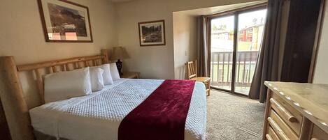 A hotel room with a bed, a wooden headboard, a nightstand, a lamp, a chair, a desk, and a view of the outside through a sliding glass door.