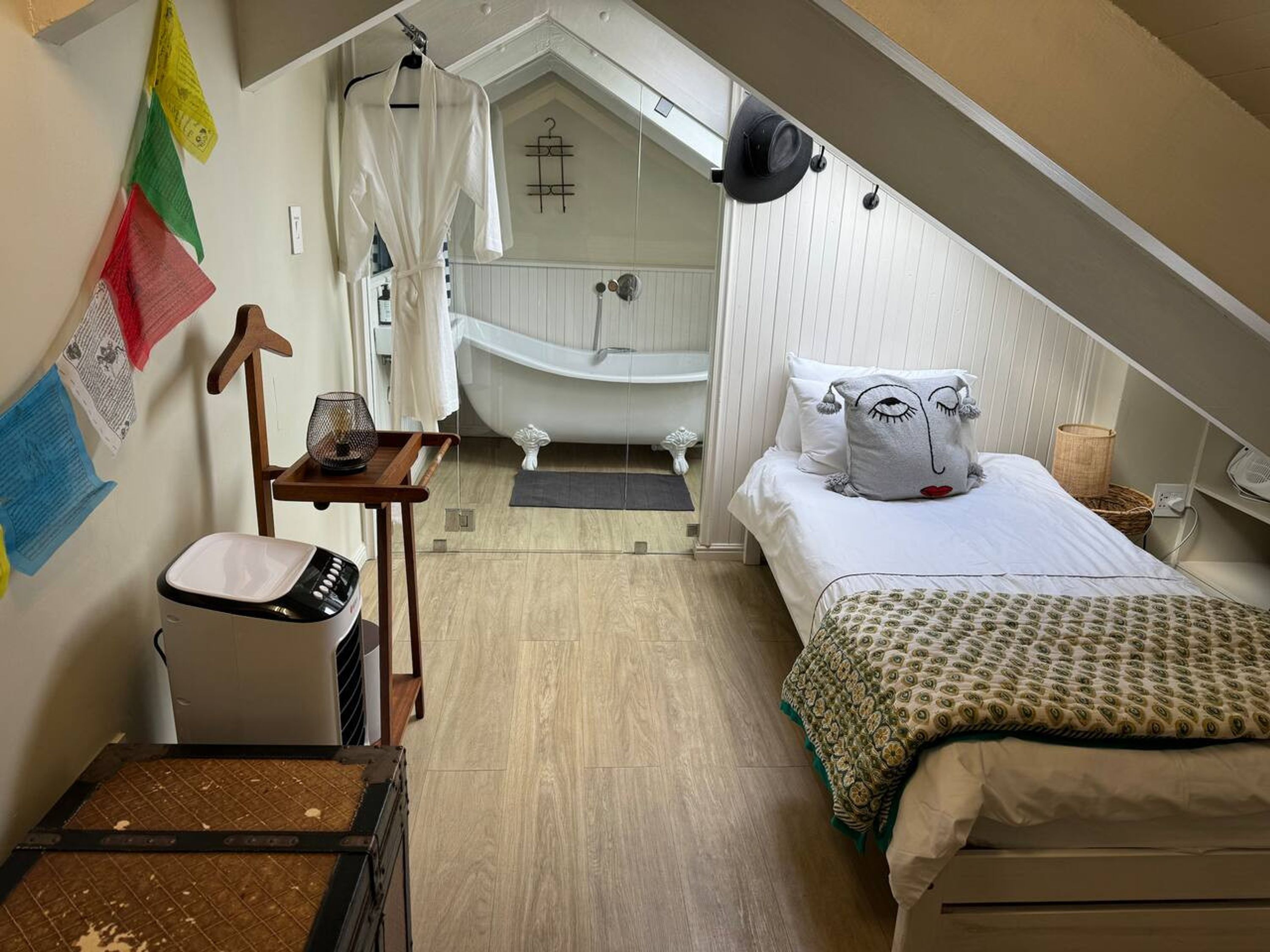 A bedroom with a bed, a small table, a chair, a suitcase, a hanging hat, and a bathtub in the background.