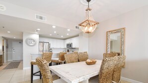 A dining area with a white table, wicker chairs, a chandelier, and a kitchen in the background. - 3rd Floor Condo w/ Large Community Pool in Central Destin! 🌺🐬 (Destin)