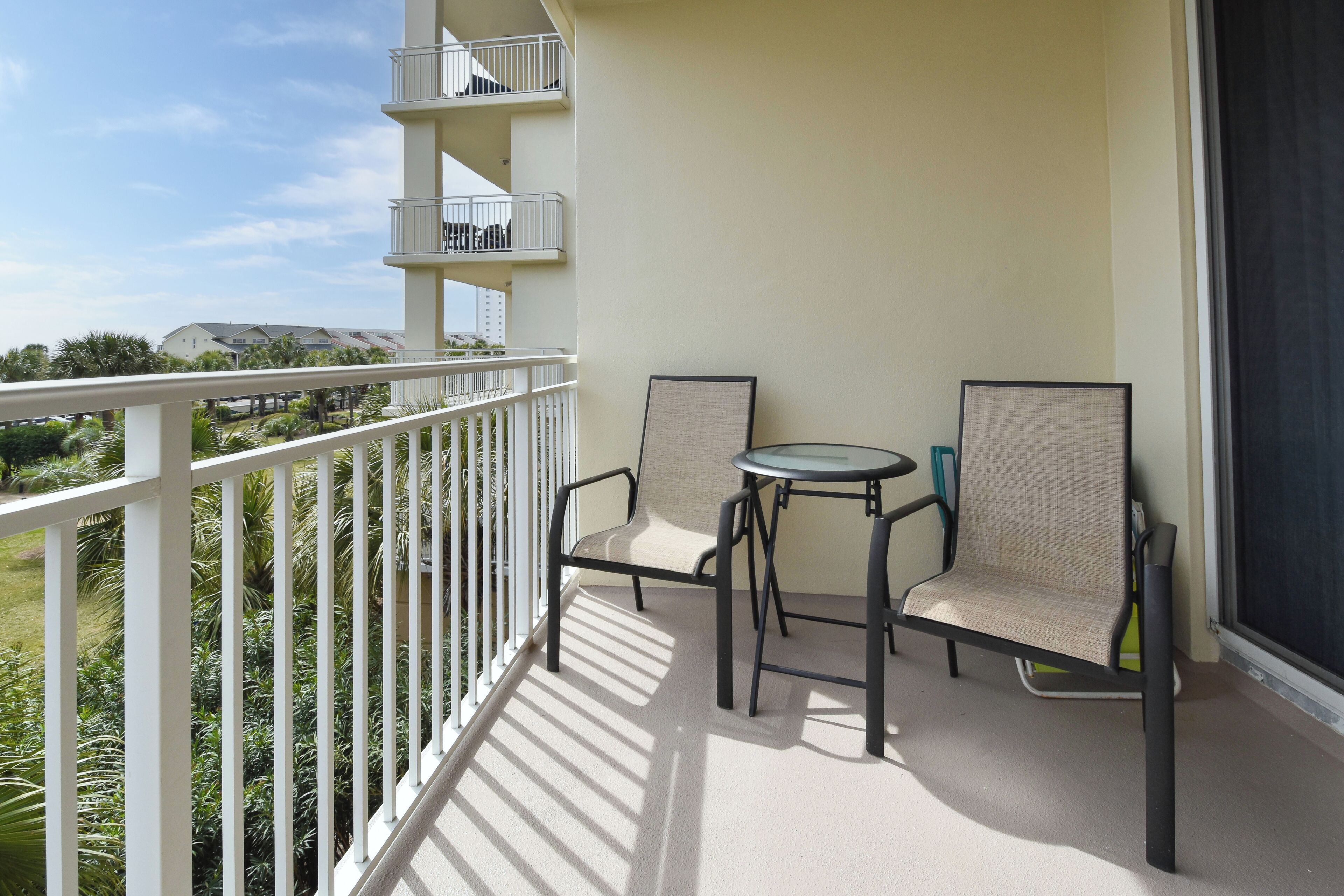Un balcon avec une balustrade blanche, deux chaises, une petite table ronde et une vue sur la verdure et les bâtiments en arrière-plan.
