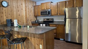 A kitchen with wooden cabinets, a stainless steel refrigerator, a microwave, an oven, and a bar stool. - Unit 624 - Updated Condo (Winter Park)