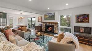 A spacious living room with a stone fireplace, comfortable seating, and large windows offering a view of the outdoors.