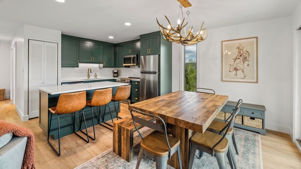 A modern kitchen with dark green cabinets, a wooden dining table, and a chandelier.