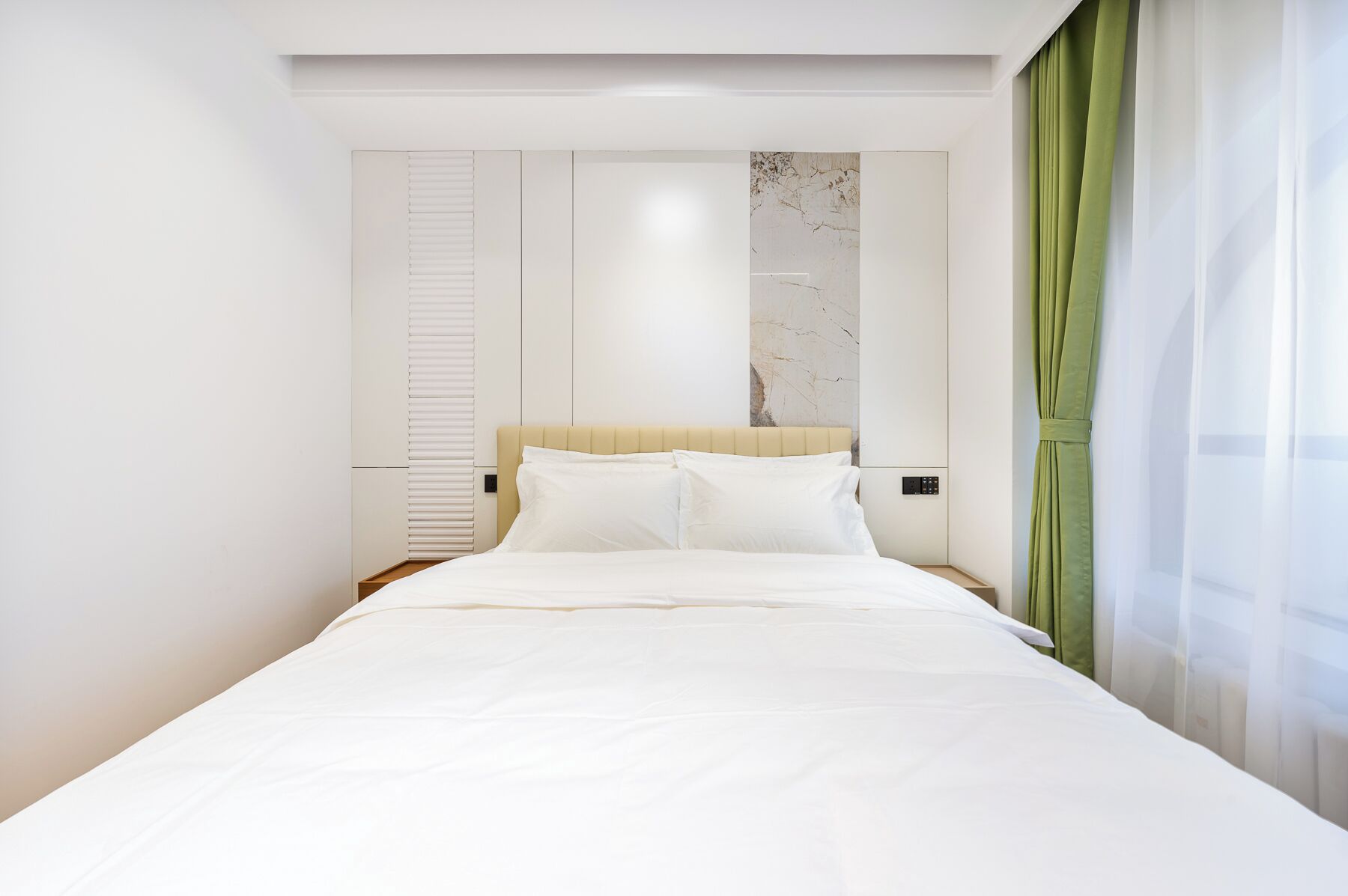 Una habitación de hotel moderna con una cama grande, un cabecero con detalles de mármol, un armario y una cortina verde.