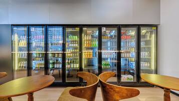 Uma área de bar moderna com um grande expositor refrigerado repleto de diversas bebidas. Cadeiras e mesas de madeira estão em primeiro plano.