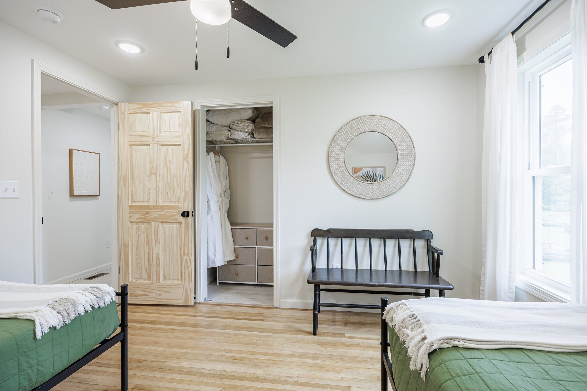 A bedroom with two beds, a wooden wardrobe, a bench, a mirror, and a window.