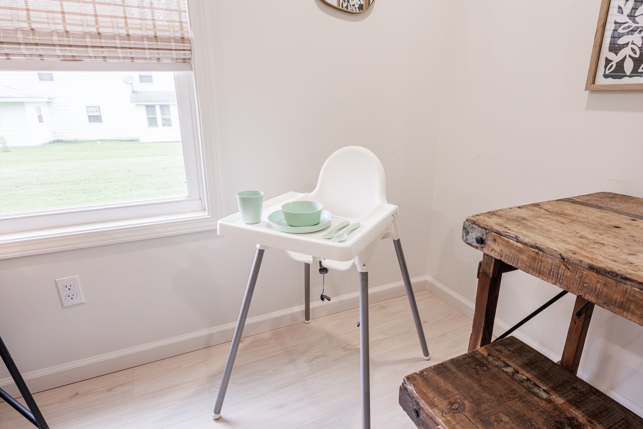 A room with a white chair with a tray, a wooden table, a window with blinds, and a power outlet on the wall.