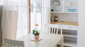 Une pièce lumineuse avec une table en bois, des chaises blanches, une plante en pot et un bureau blanc doté d’étagères.
