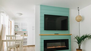 Un salon moderne avec un mur d’accent de couleur turquoise, un téléviseur à écran plat, une cheminée et un coin repas avec des chaises blanches.