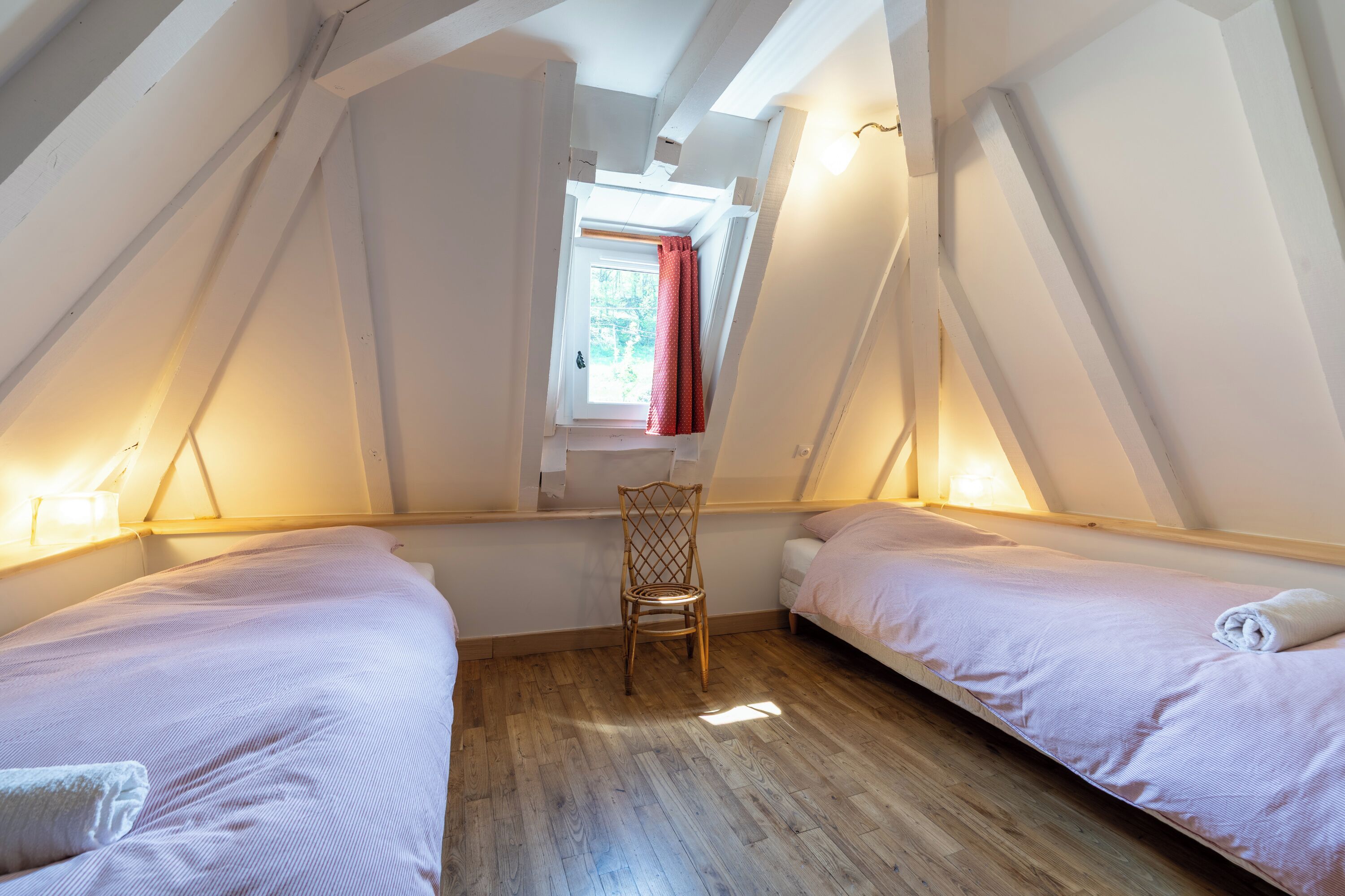 A room with two beds, a wooden floor, a window with a red curtain, and a chair.