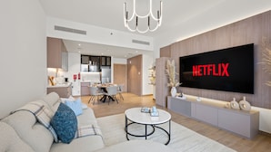 A modern living room with a large flat-screen TV displaying 'NETFLIX', a dining area, and a kitchen.