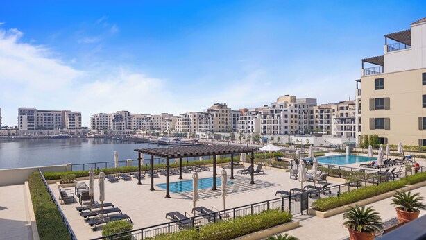 A rooftop pool area with lounge chairs and umbrellas, overlooking a marina with boats and a cityscape in the background.