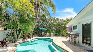 A swimming pool with a curved edge, surrounded by palm trees and a patio area with outdoor furniture.