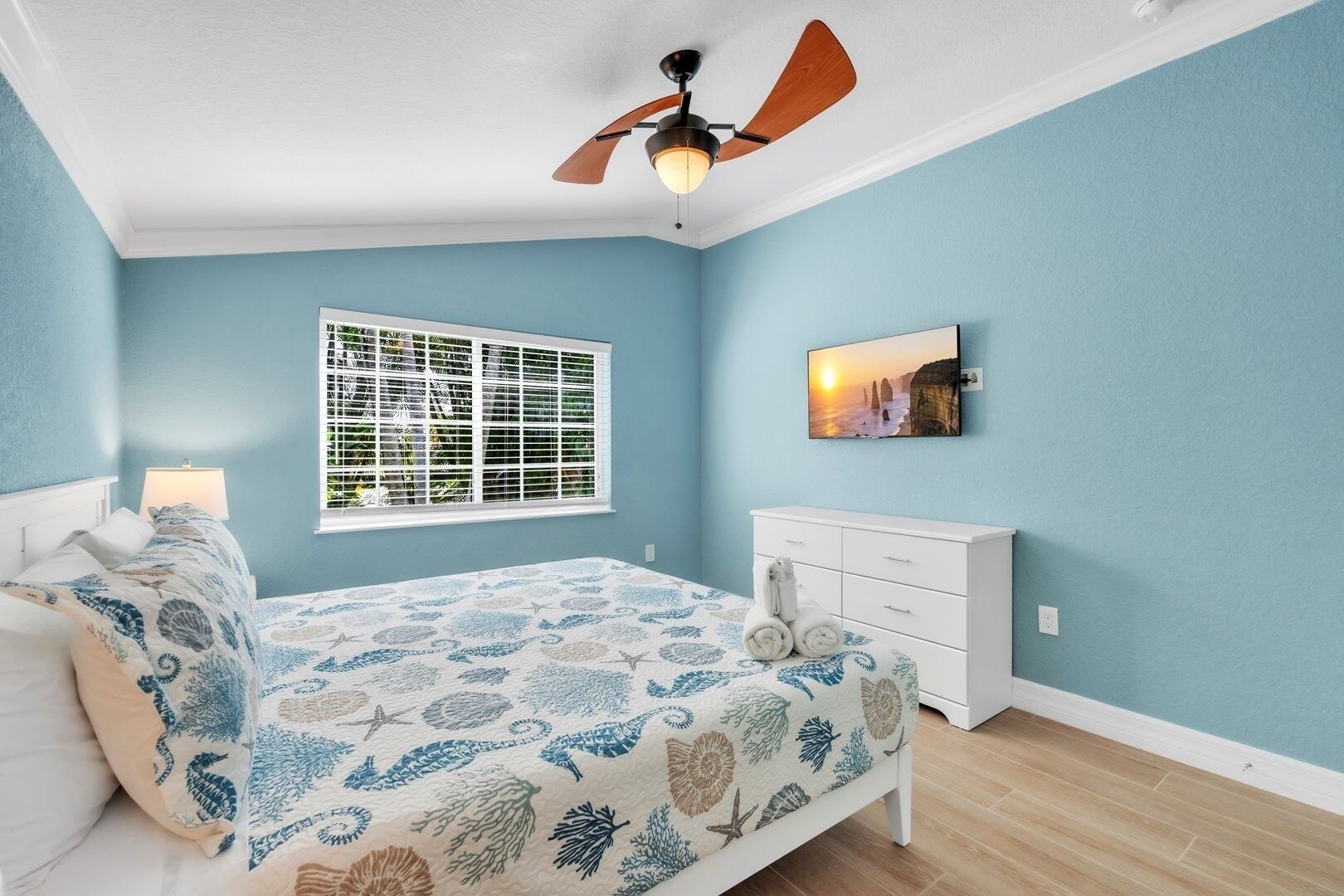 A bedroom with a bed, a ceiling fan, a window with blinds, a dresser, and a wall-mounted TV.