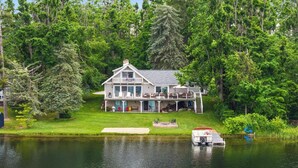 A two-story house with a deck, a boat docked by the water, and a lawn.