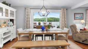 A dining area with a wooden table, chairs, a bench, a white cabinet with glass doors, a chandelier, and a view of a lake through a glass door.