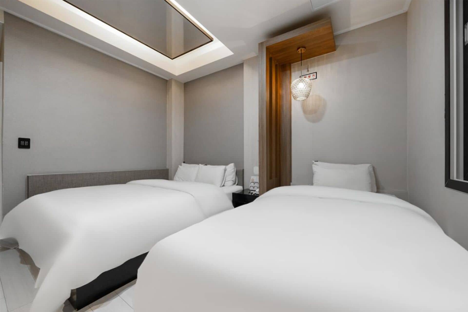 A hotel room with two beds, a skylight, a wooden accent wall, and a hanging light fixture.