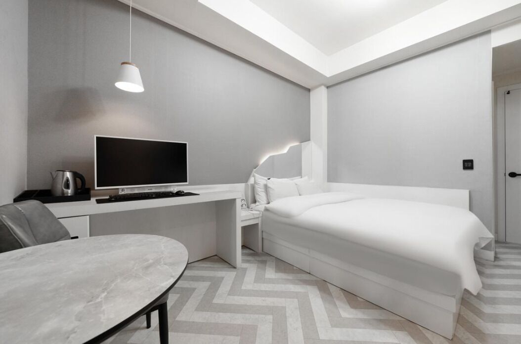 A modern hotel room with a bed, a desk with a television, a chair, and a table.